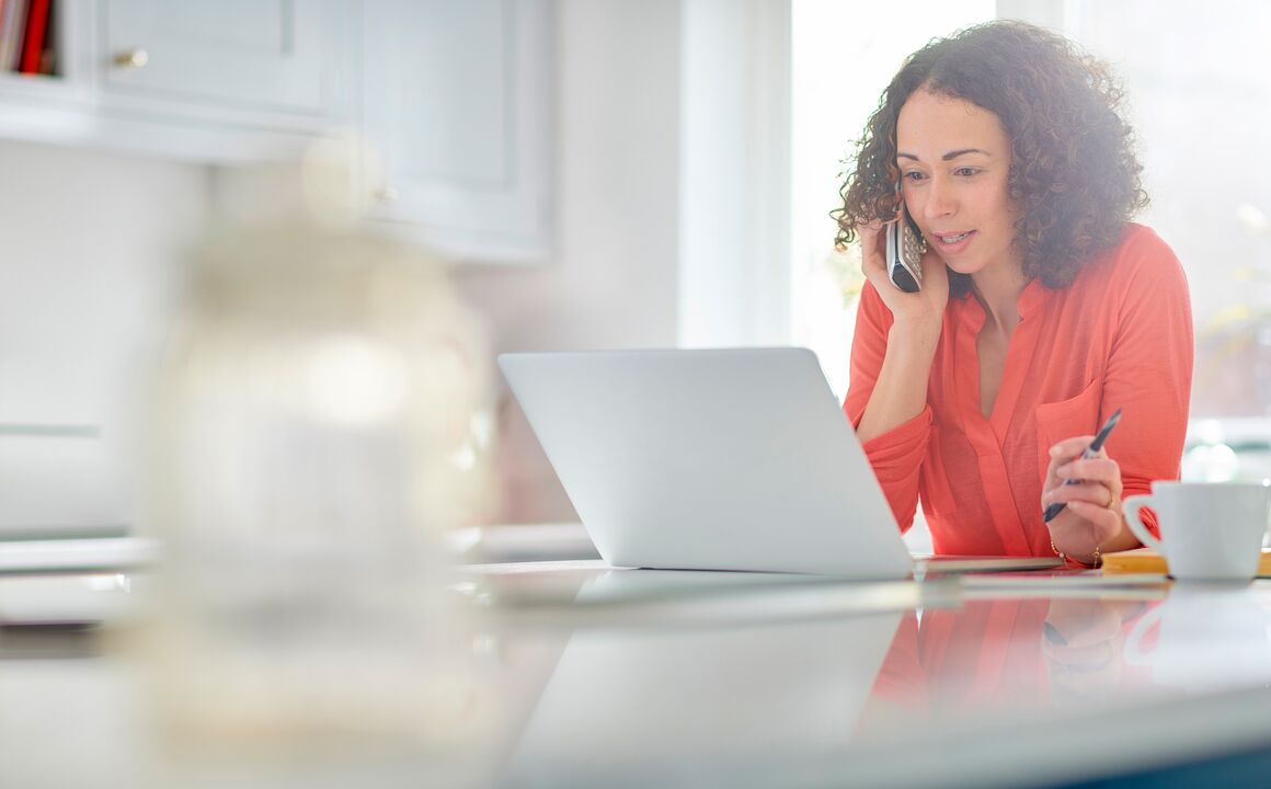 Woman sits in her kitchen while she looks at a laptop screen and speaks to a health insurance exchange customer service representative