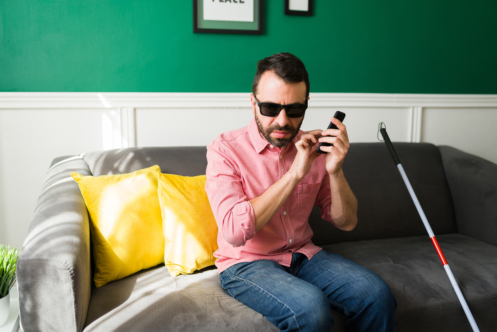 Blind man with a cane sits on a couch and holds up a phone in one hand while tapping the screen with the other hand. 