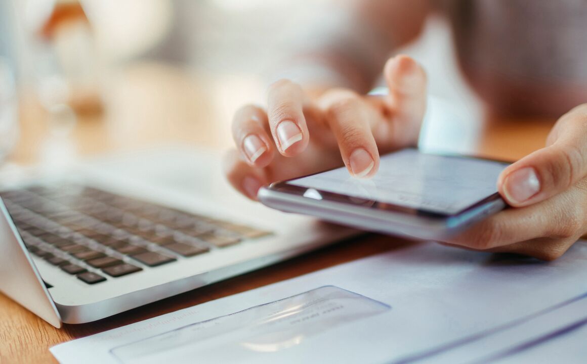 Close-up of hands holding a mobile device being used to select a health plan. An open laptop and a stack of mail are nearby .  